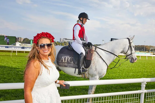 A woman in a white dress and red flower crown smiles by a horse rider wearing a red vest at a racetrack.