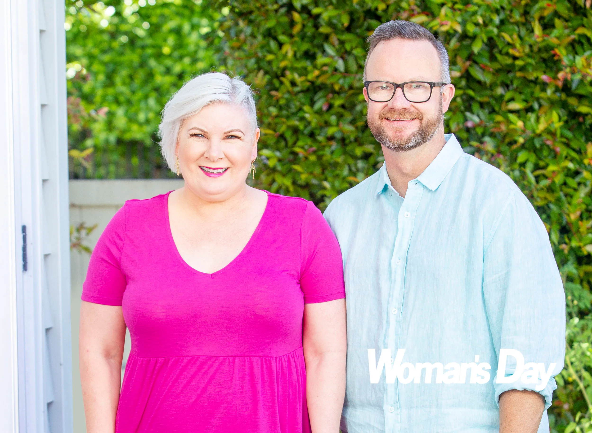 A woman in a pink top stands beside a man in a light blue shirt. They are outdoors with greenery behind them.