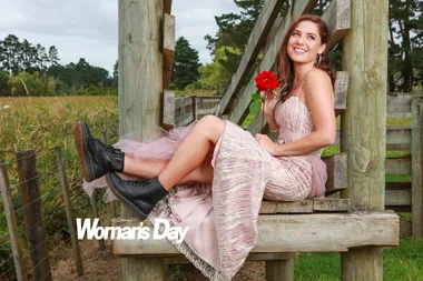 A woman in a pink dress and black boots sits on a wooden structure holding a red rose, smiling outdoors.