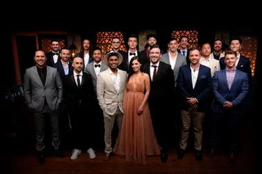 A woman in a peach gown stands with a group of well-dressed men, posing in front of a decorative background.