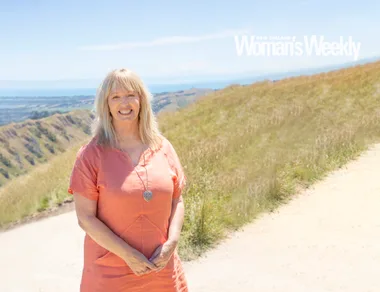 A woman in an orange dress stands on a scenic hillside path with a view of the ocean and hills in the background.
