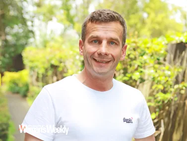 A man in a white T-shirt smiles outdoors with greenery in the background. New Zealand Woman's Weekly logo is visible.