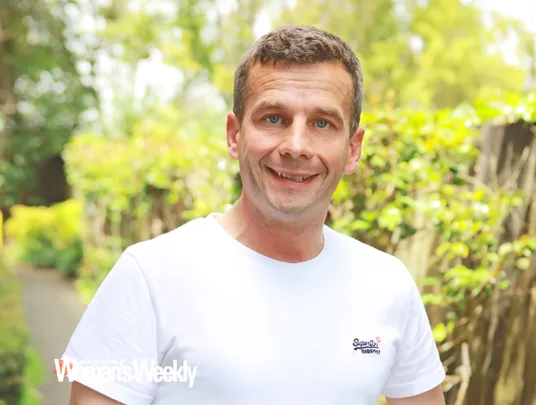 A man in a white T-shirt smiles outdoors with greenery in the background. New Zealand Woman's Weekly logo is visible.