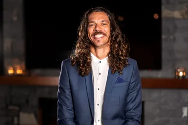 Man with long curly hair and a mustache smiling, wearing a blue suit and white shirt, in dimly lit room.