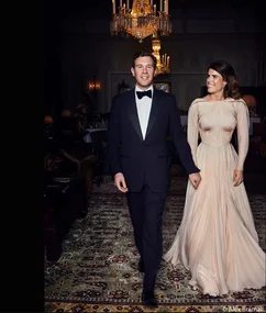 Man in tuxedo and woman in elegant gown walking together in a luxurious room under a chandelier.