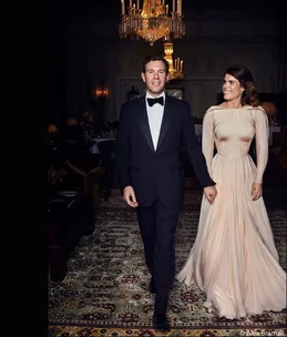 Man in tuxedo and woman in elegant gown walking together in a luxurious room under a chandelier.