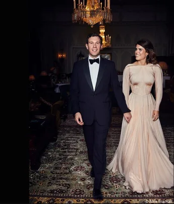 Man in tuxedo and woman in elegant gown walking together in a luxurious room under a chandelier.