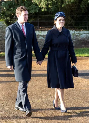 Man and woman walking hand in hand in matching dark coats, with the woman wearing a blue headpiece.