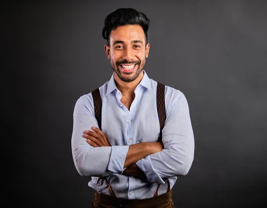 Smiling man in a light blue shirt with suspenders, arms crossed, against a dark gray background.