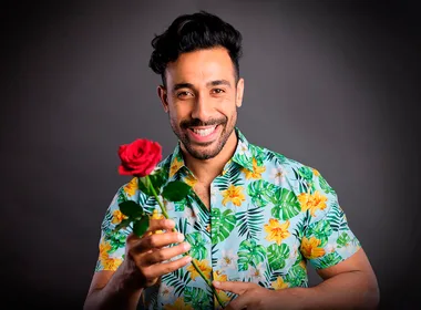 Man in a colorful floral shirt smiling and holding a red rose against a dark background.