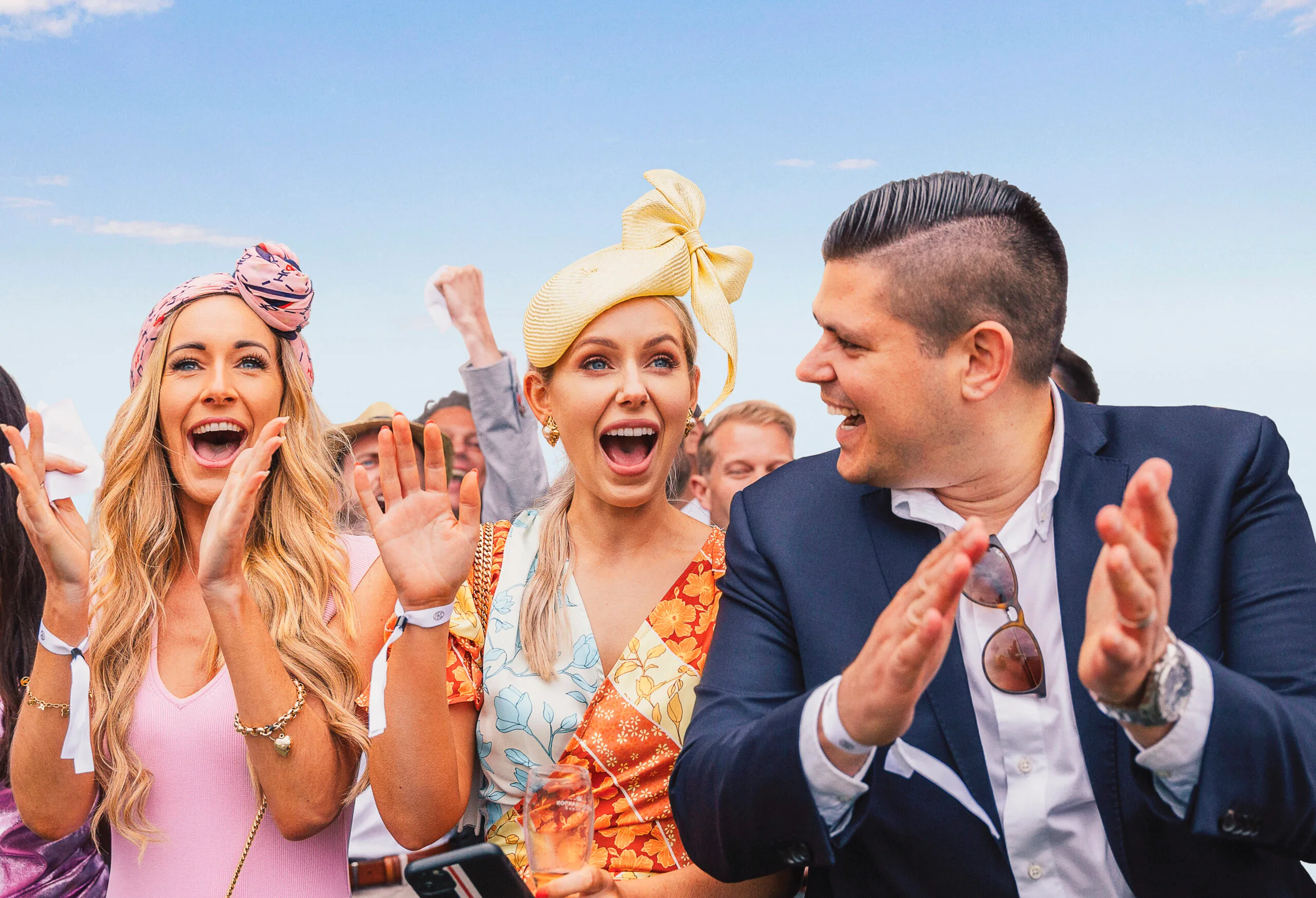 Three people at an outdoor event smiling and clapping, wearing hats and formal attire.