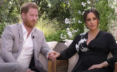 A couple holding hands, seated outdoors on wooden chairs, surrounded by greenery and white flowers.