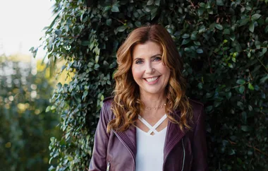 Smiling woman in a leather jacket leans against a wall of leaves.