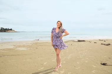 A woman in a purple floral dress standing on a sandy beach with the ocean in the background.