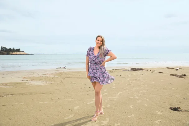 A woman in a purple floral dress standing on a sandy beach with the ocean in the background.