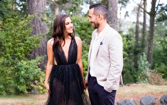 A woman in a black dress and a man in a light suit smile at each other outdoors.