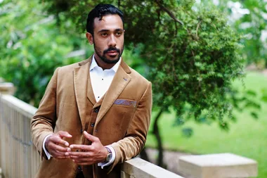 Man in a brown suit with a patterned pocket square, leaning on a railing in a green outdoor setting.