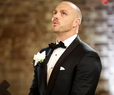 A man in a tuxedo, with a white boutonniere, looks upward against a blurred brick wall background.
