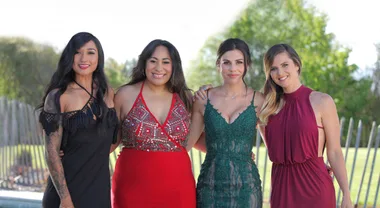 Four women in elegant dresses pose together outdoors with greenery in the background.