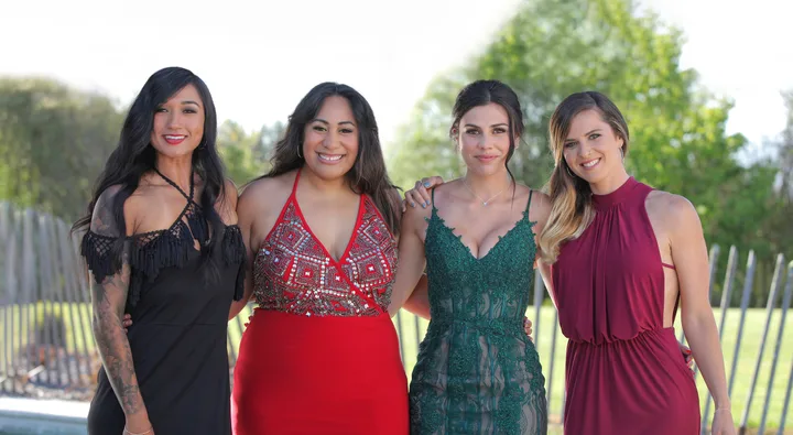 Four women in elegant dresses pose together outdoors with greenery in the background.