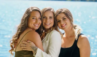 Three women smiling and posing together by a bright blue lake.