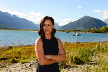 Woman with folded arms, smiling by a lake with mountains in the background.