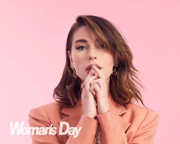 Woman in a peach blazer posing with hands near face against a pink background, wearing gold earrings; text: Woman's Day.