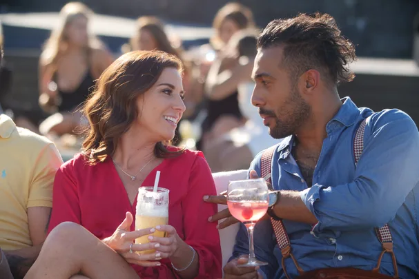 A woman in a red dress and a man in a blue shirt converse, holding drinks outdoors with people in the background.
