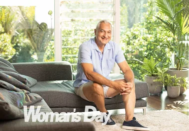 Man in light-blue shirt sitting on a sofa in a sunlit room with plants. "Woman's Day" logo in the corner.
