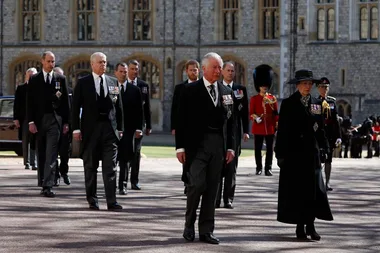 Royal family members in formal attire walk in a procession outside a historic stone building.