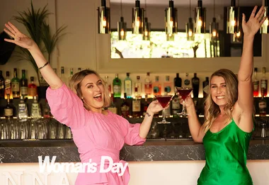 Two women in a bar, smiling and holding cocktails, celebrate with arms raised.