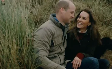 A smiling couple sitting together in tall grass, wearing casual jackets, looking at each other.