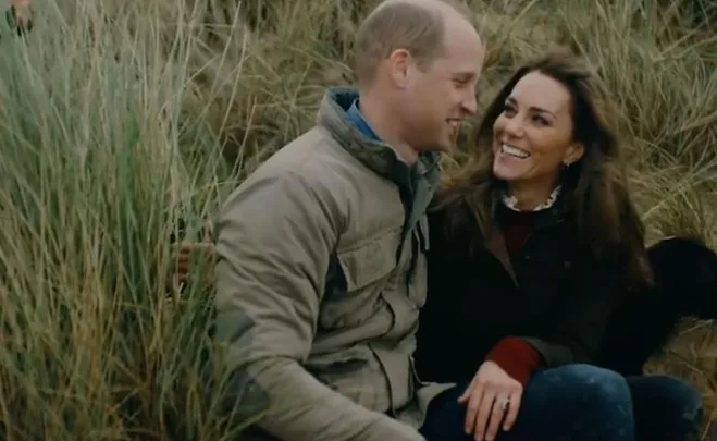 A smiling couple sitting together in tall grass, wearing casual jackets, looking at each other.