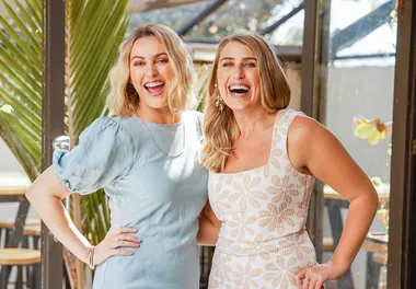 Two women smiling, one in a light blue dress and the other in a floral white dress, standing indoors.
