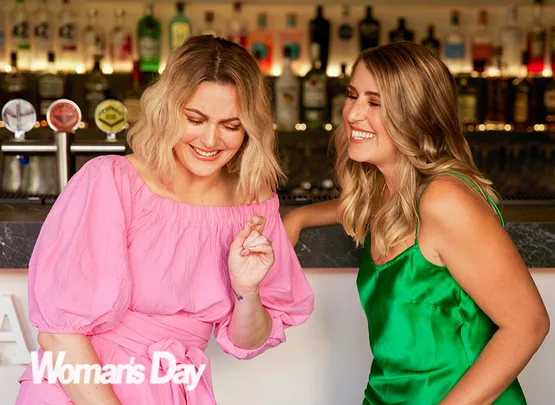 Two women laughing together at a bar, one in a pink dress and the other in a green dress, with a "Woman's Day" logo.