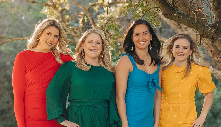 Four women in colorful dresses standing outdoors, smiling at the camera amidst trees and sunlight.