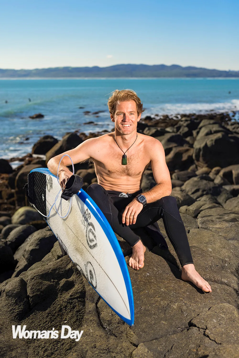 Billy Stairmand sitting on a rock with his wetsuit around his waist, holding his surfboard
