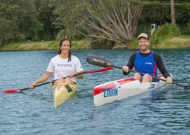 Two people kayaking on a calm lake, smiling, with greenery in the background.