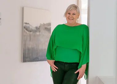 A woman with short blonde hair in a green blouse leans against a white wall in a bright room.