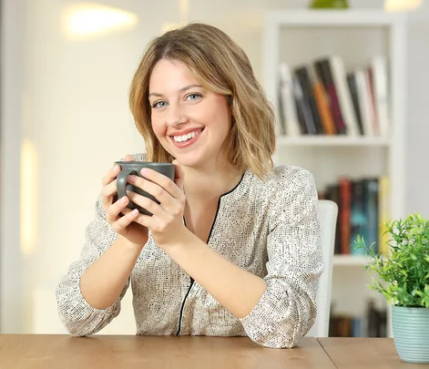 Smiling person holding a mug at a wooden table with a bookshelf in the background.