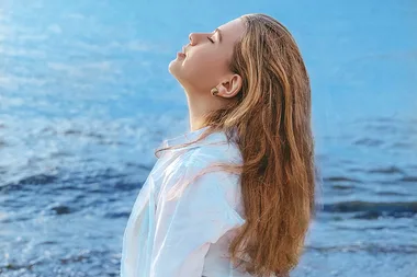 Profile of a woman with long hair by the sea, wearing a white shirt, looking upwards with eyes closed in relaxation.