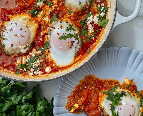 Shakshuka with poached eggs, tomatoes, herbs, and spices in a skillet, served with parsley garnish.