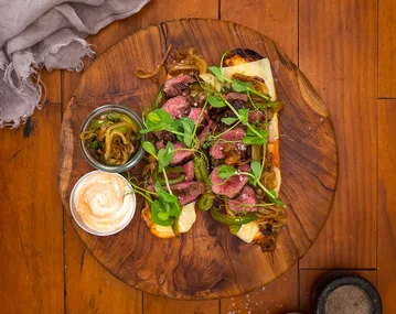 Grilled steak flatbread with caramelized onions, peppers, greens, and sauces on a wooden board.
