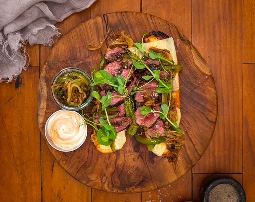 Grilled steak flatbread with caramelized onions, peppers, greens, and sauces on a wooden board.