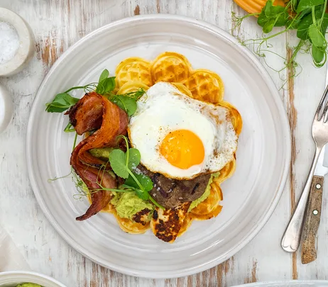 Waffles topped with fried egg, bacon, and greens on a white plate set on a rustic wooden table.