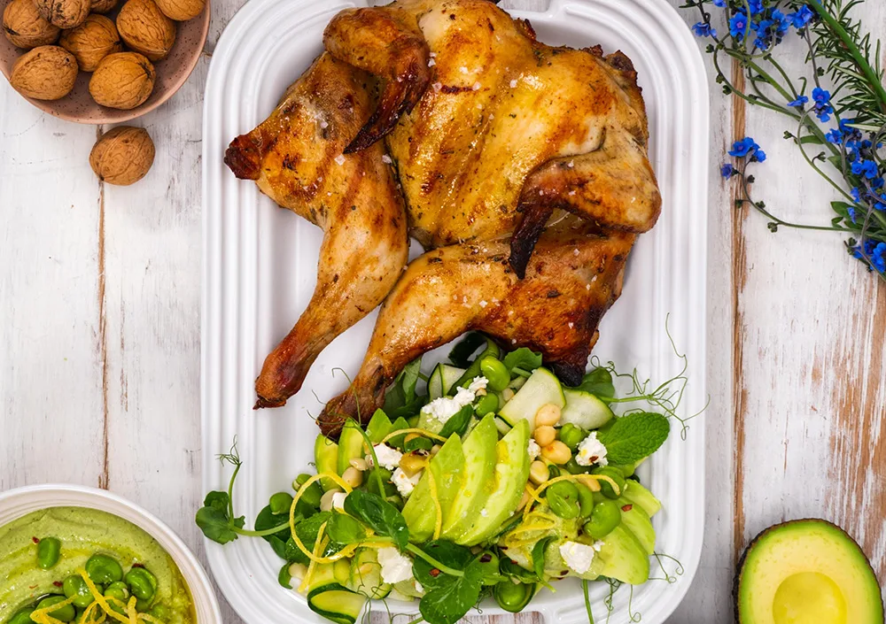 Roasted chicken with a fresh green salad and avocados on a white platter, surrounded by nuts and blue flowers.