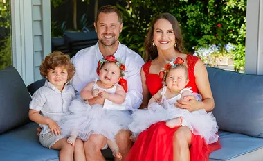A smiling family of four, with two babies in white outfits, seated on a couch outdoors.