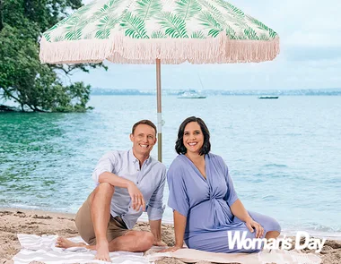 A man and a woman sitting on a beach under a decorative umbrella, smiling, with the sea in the background.