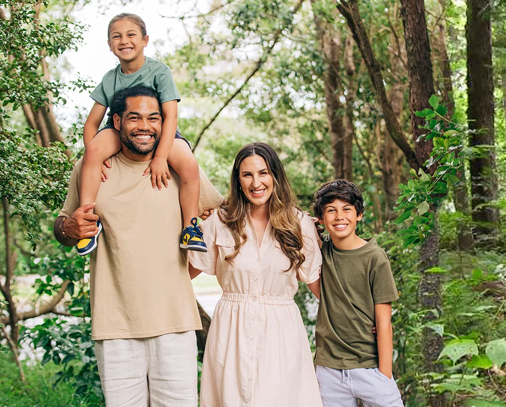 Family of four smiling in a lush green forest, with a man carrying a child on his shoulders.