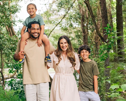 Family of four smiling in a lush green forest, with a man carrying a child on his shoulders.